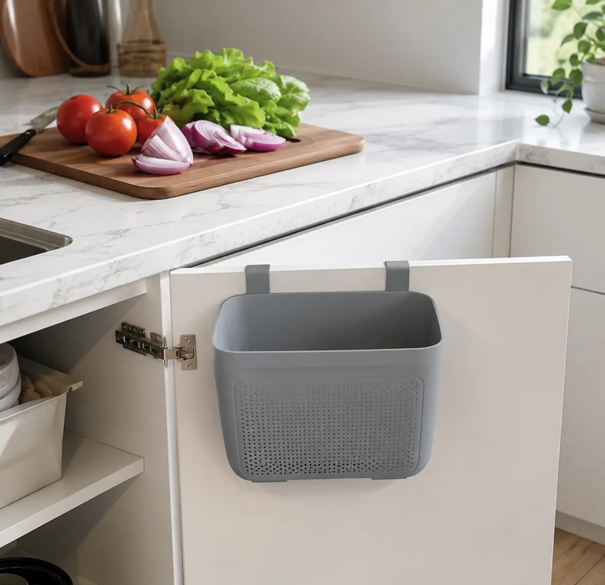Mini cubo de basura plegable gris colgado en la puerta de un armario de cocina, con verduras frescas visibles sobre la encimera